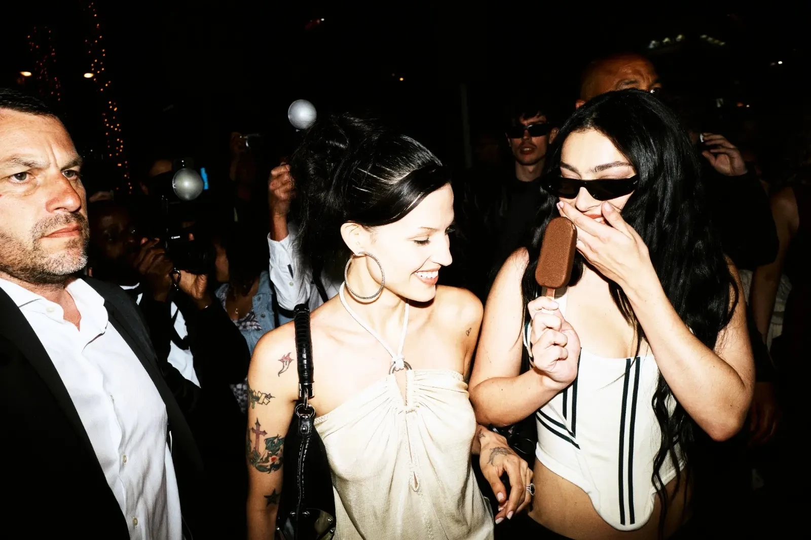 Smiling young woman with hoop earrings and arm tattoos beside a friend holding a chocolate ice cream bar.