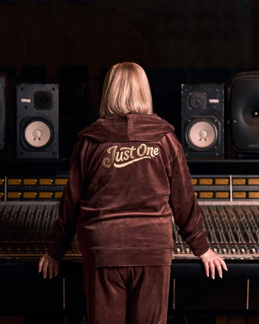 Woman seen from behind at a studio mixing console, wearing a brown velour hoodie embroidered with “Just One.”