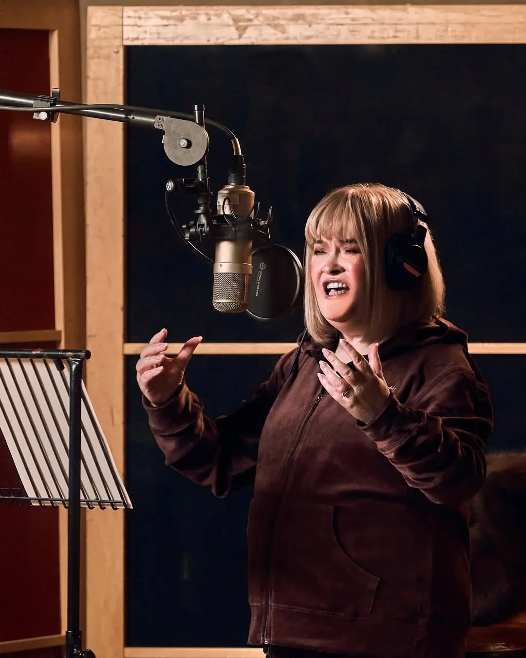 Woman in headphones sings passionately into a studio microphone, wearing a brown velour hoodie.