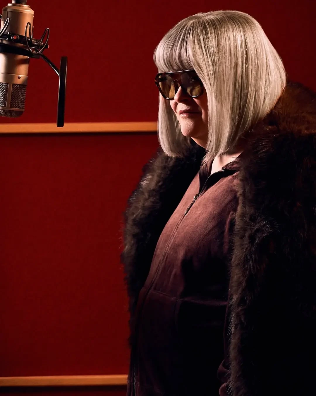 Woman with a platinum bob, tinted sunglasses and a fur coat poses beside a microphone in a red recording studio.