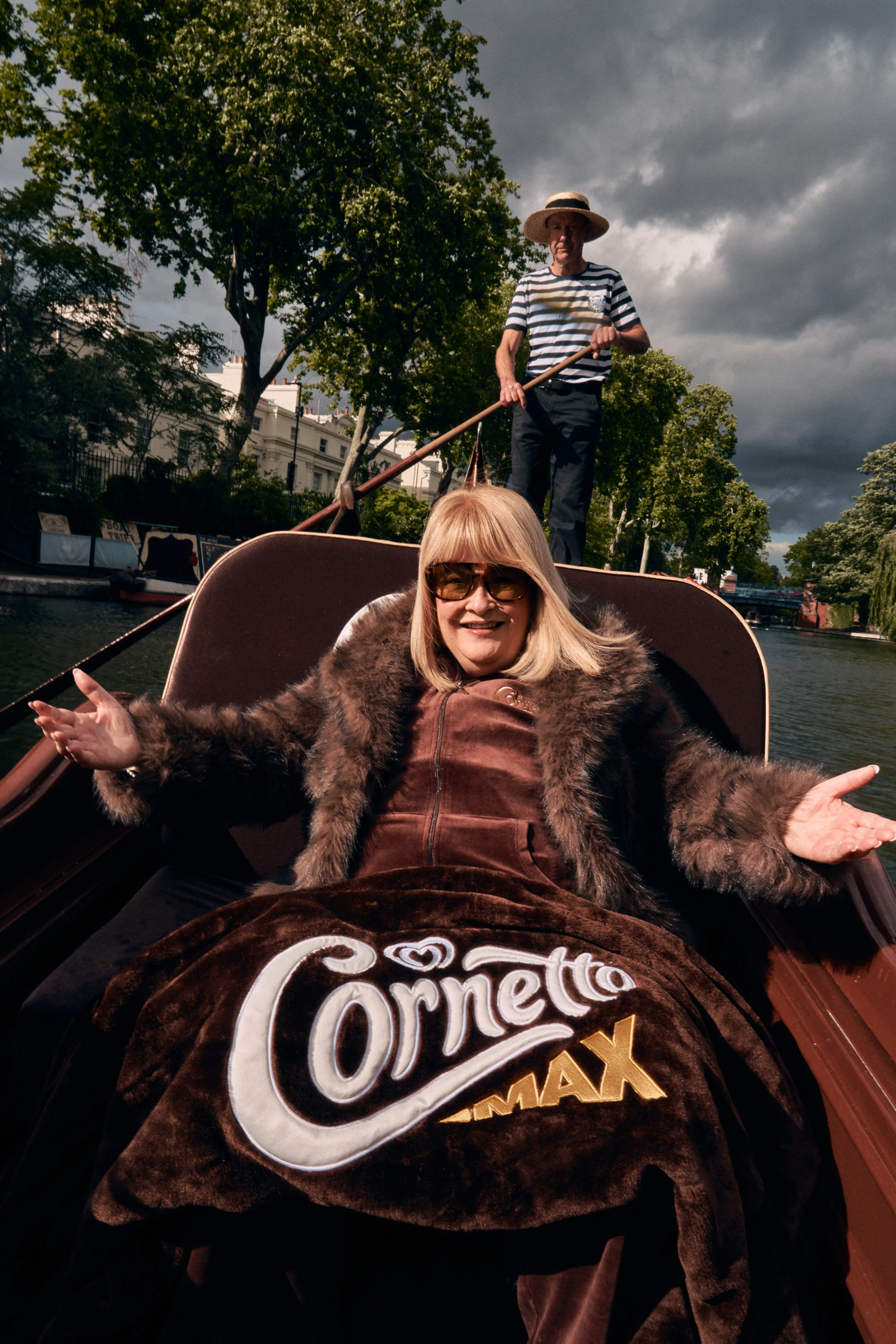 Smiling woman in sunglasses and a fur coat rides a gondola on a canal, draped in a Cornetto Max blanket.